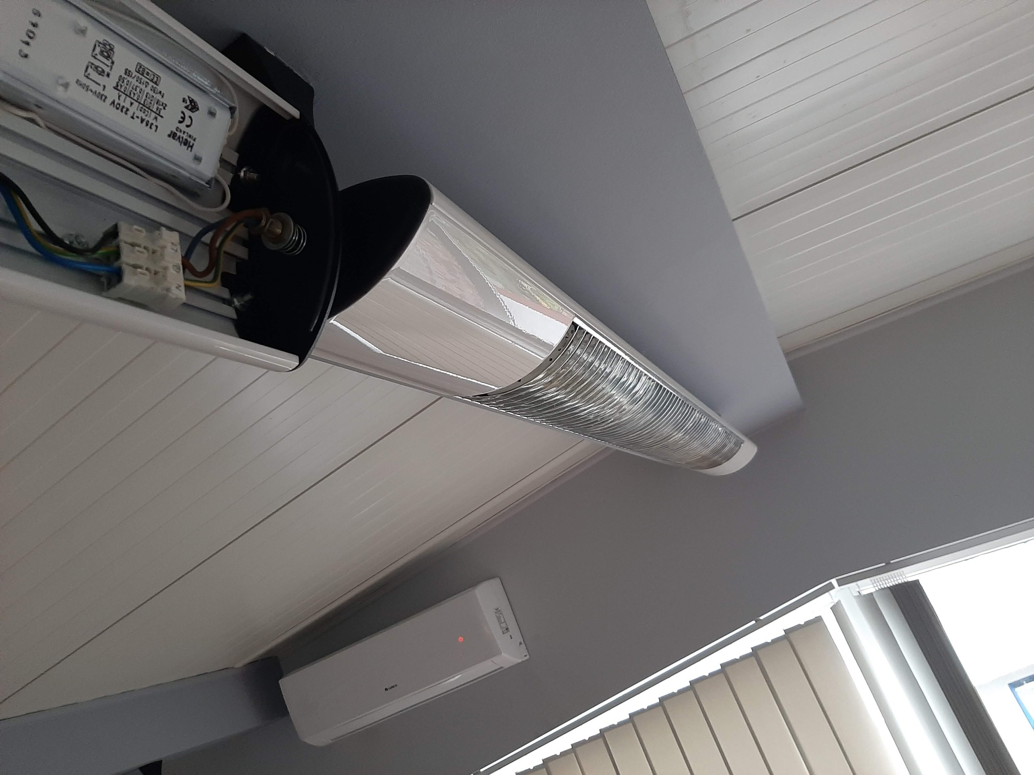 Otwarta obudowa lampy LED na suficie z widocznymi elementami elektrycznymi i złączkami, obok klimatyzator i żaluzje.