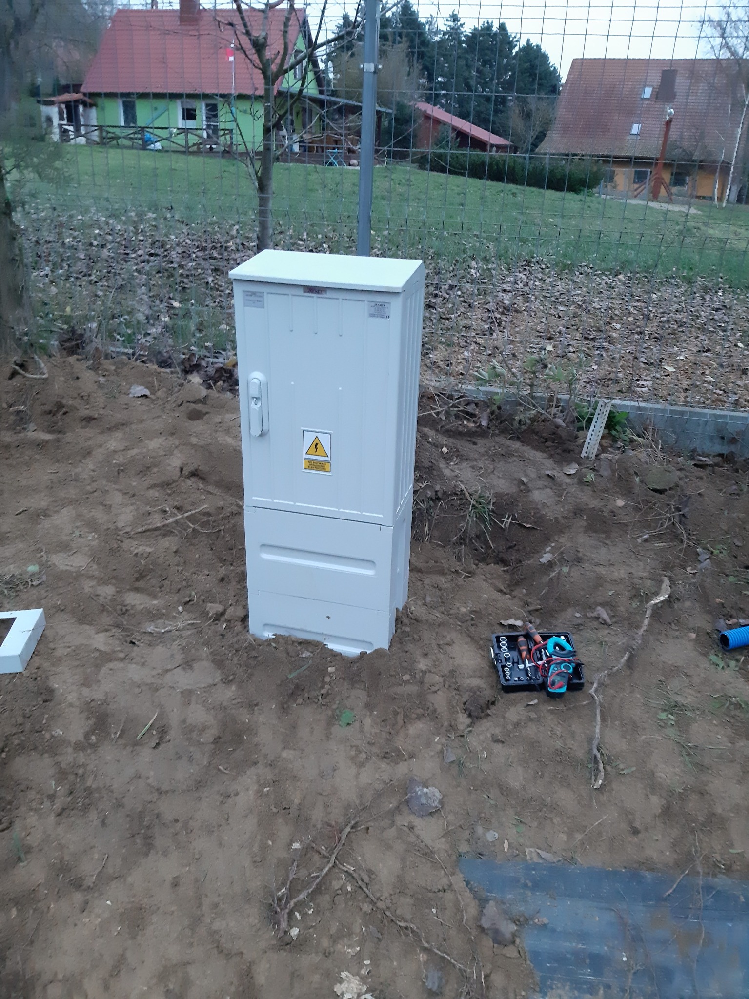 Nowa, biała skrzynka zasilania zewnętrznego, z naklejką ostrzegawczą o wysokim napięciu, umieszczona w wykopie, obok leży czarna skrzynka z narzędziami elektrycznymi i niebieska rura karbowana.