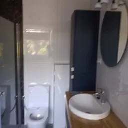 Nowoczesna łazienka z białą ceramiką, umywalką nablatową na drewnianym blacie i ciemnoniebieską szafką, widoczna kabina prysznicowa z ciemną ramą.