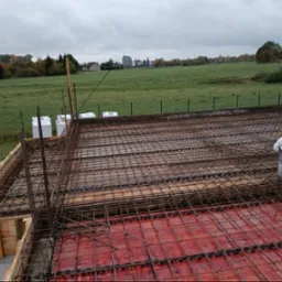 Zbrojenie stropu na czerwonym szalunku z widokiem na zielone pola i zabudowania w tle, pochmurny dzień.