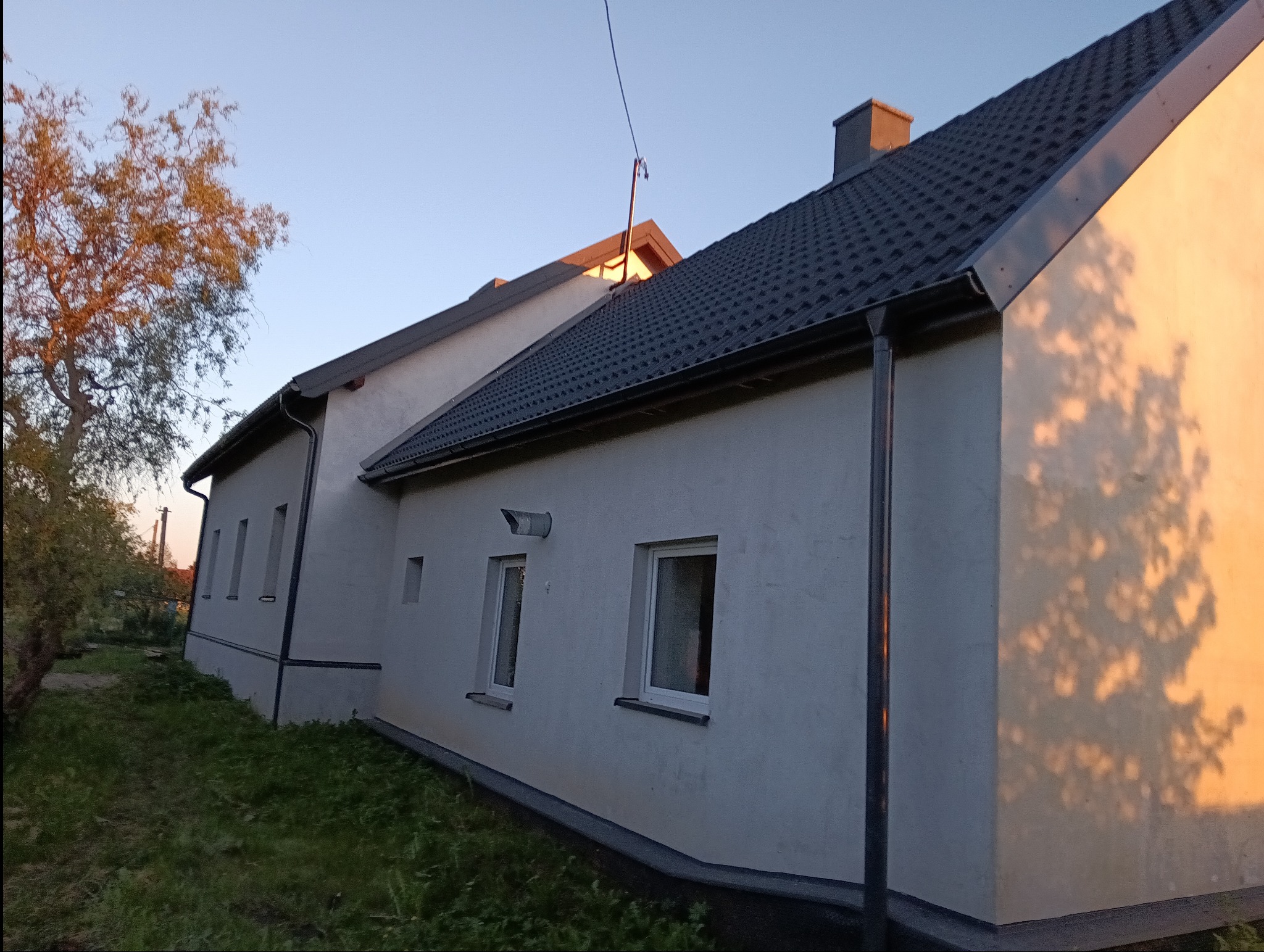 Zewnętrzny widok boczny domu z szarą dachówką, białymi ścianami i widocznymi oknami, oświetlony ciepłym światłem zachodzącego słońca, rzucającym cienie drzew na elewację.