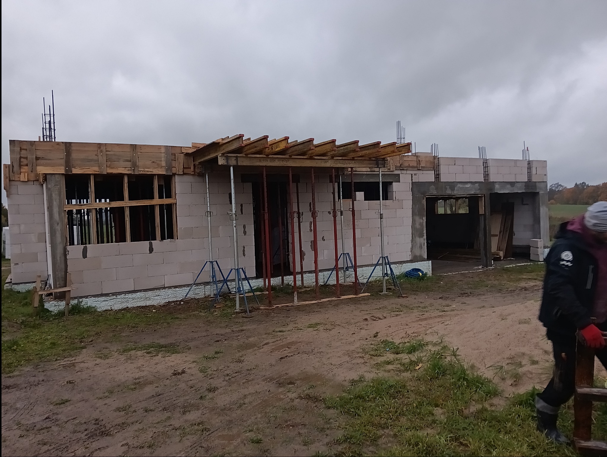 Budowa domu z bloczków betonowych w trakcie realizacji, widoczne szalunki drewniane, podpory stalowe i zbrojenia, na pierwszym planie pracownik w odzieży roboczej.