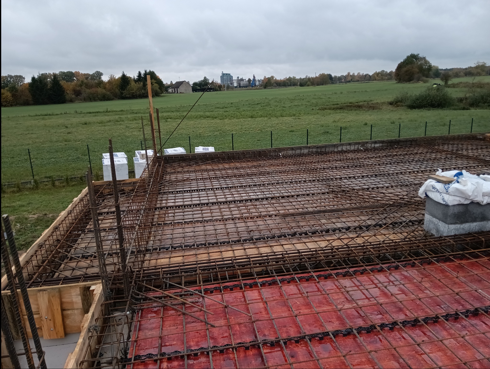 Zbrojenie stropu na czerwonym szalunku z widokiem na zielone pola i zabudowania w tle, pochmurny dzień.
