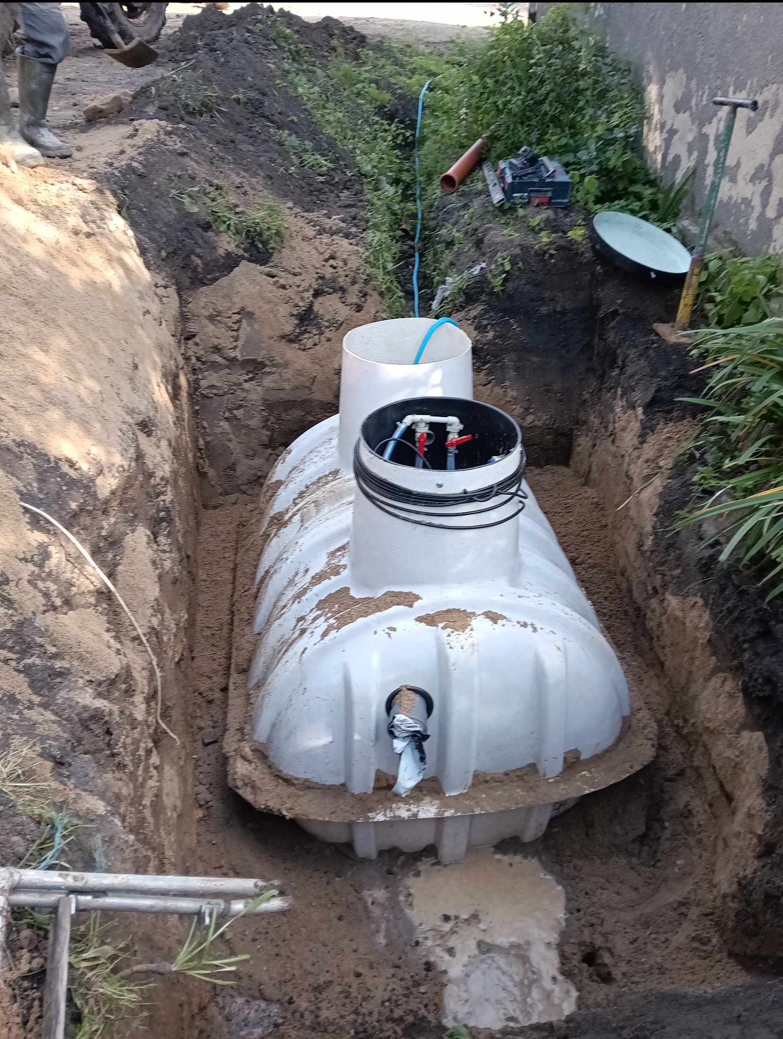 Montaż szamba betonowego w wykopie, widoczne przyłącza i elementy instalacji, częściowo zasypane piaskiem, narzędzia i sprzęt budowlany w tle.