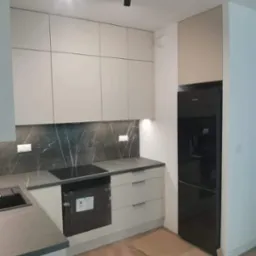 Nowoczesna kuchnia z beżowymi frontami szafek, blatem imitującym marmur, czarną płytą indukcyjną i lodówką, oświetleniem szynowym i kartonami na podłodze sugerującymi prace wykończeniowe.