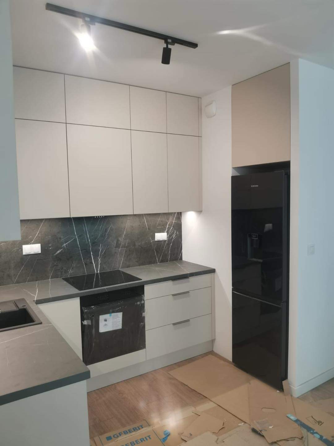 Nowoczesna kuchnia z beżowymi frontami szafek, blatem imitującym marmur, czarną płytą indukcyjną i lodówką, oświetleniem szynowym i kartonami na podłodze sugerującymi prace wykończeniowe.