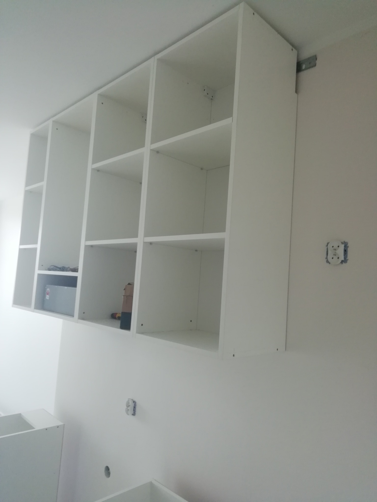 Biała, wisząca szafka kuchenna z otwartymi półkami, zamontowana na ścianie z widocznym gniazdkiem elektrycznym i elementami montażowymi, w trakcie instalacji.