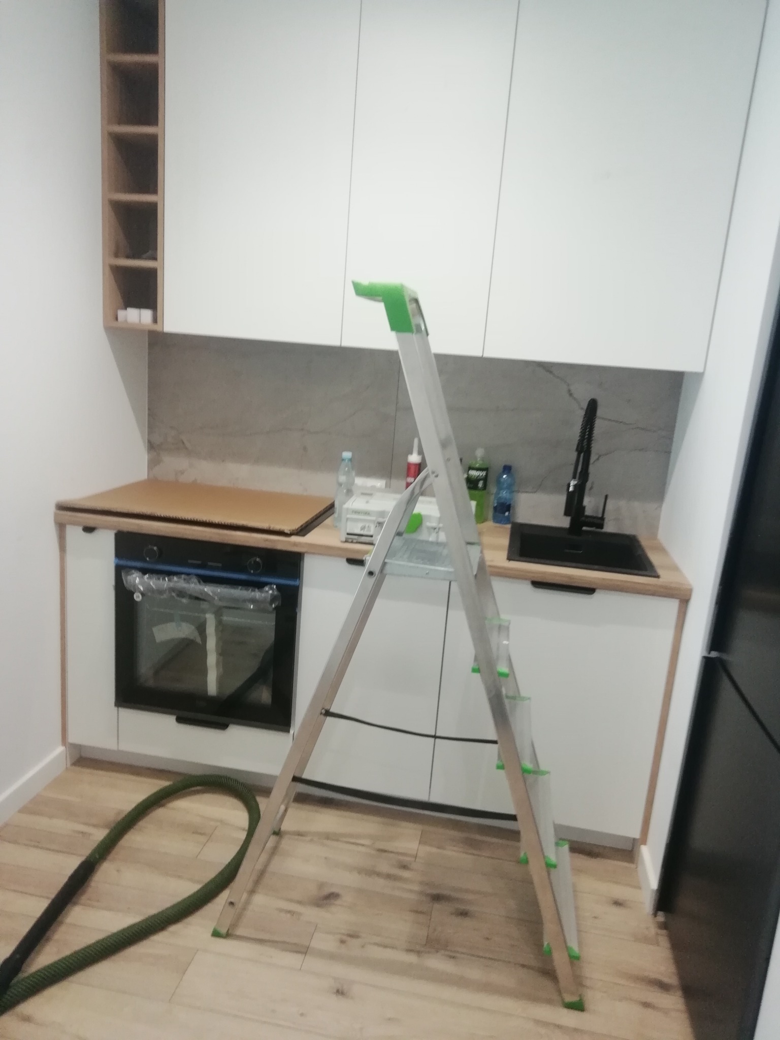 Montaż białej kuchni z szarym blatem: widoczna drabina, elementy instalacji i akcesoria, kartonowe zabezpieczenie blatu nad piekarnikiem oraz rura odkurzacza na podłodze.