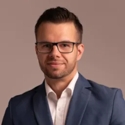 Portret młodego mężczyzny w okularach i niebieskiej marynarce, z delikatnym uśmiechem, na jednolitym, stonowanym tle.