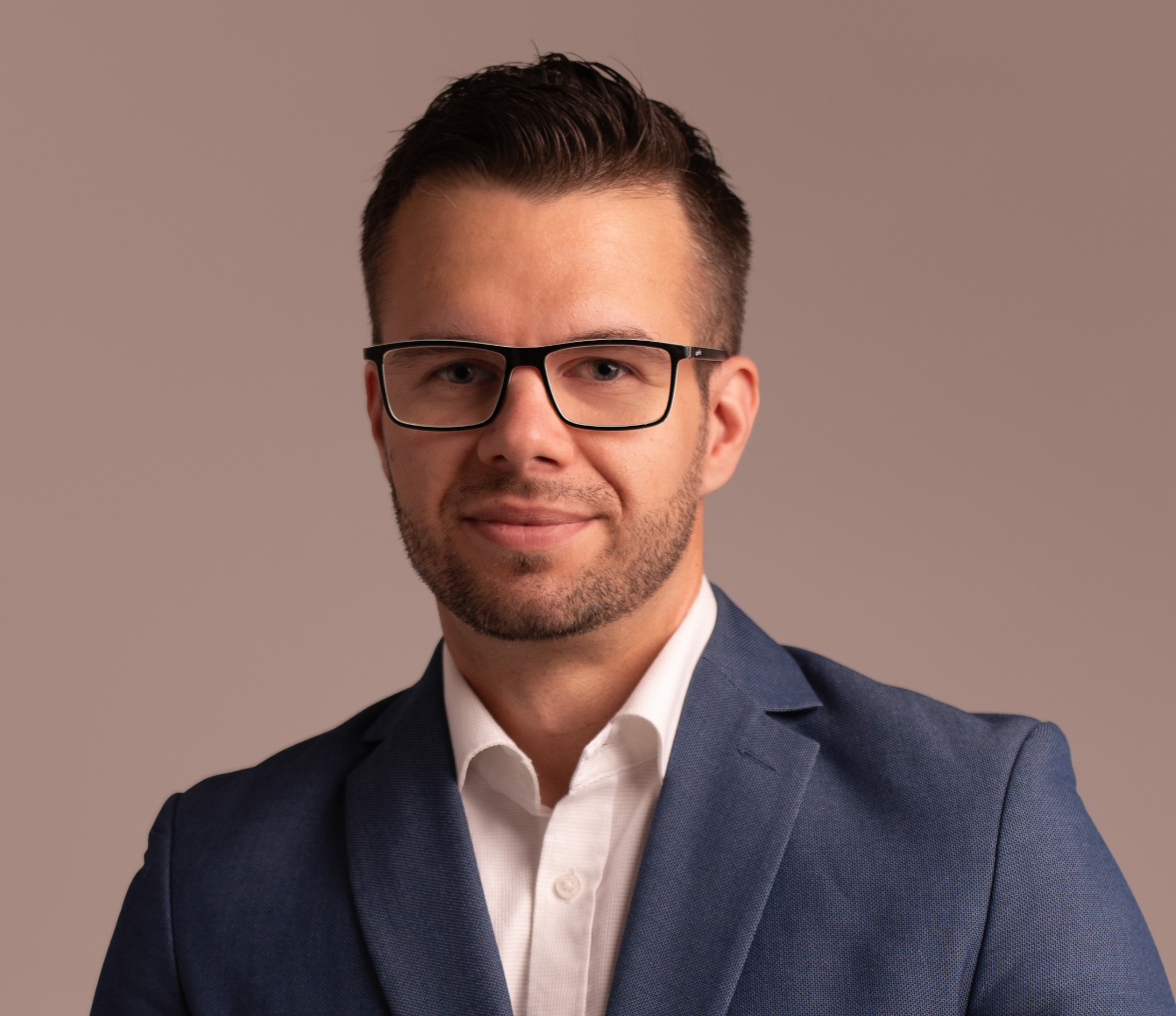 Portret młodego mężczyzny w okularach i niebieskiej marynarce, z delikatnym uśmiechem, na jednolitym, stonowanym tle.