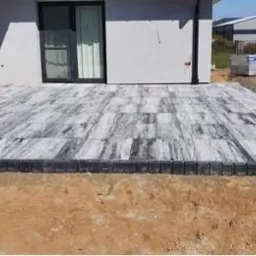 Ułożona szaro-biała kostka brukowa przed nowoczesnym domem z czarnymi ramami okiennymi, obrzeża z ciemnej kostki, teren wokół w trakcie przygotowania.