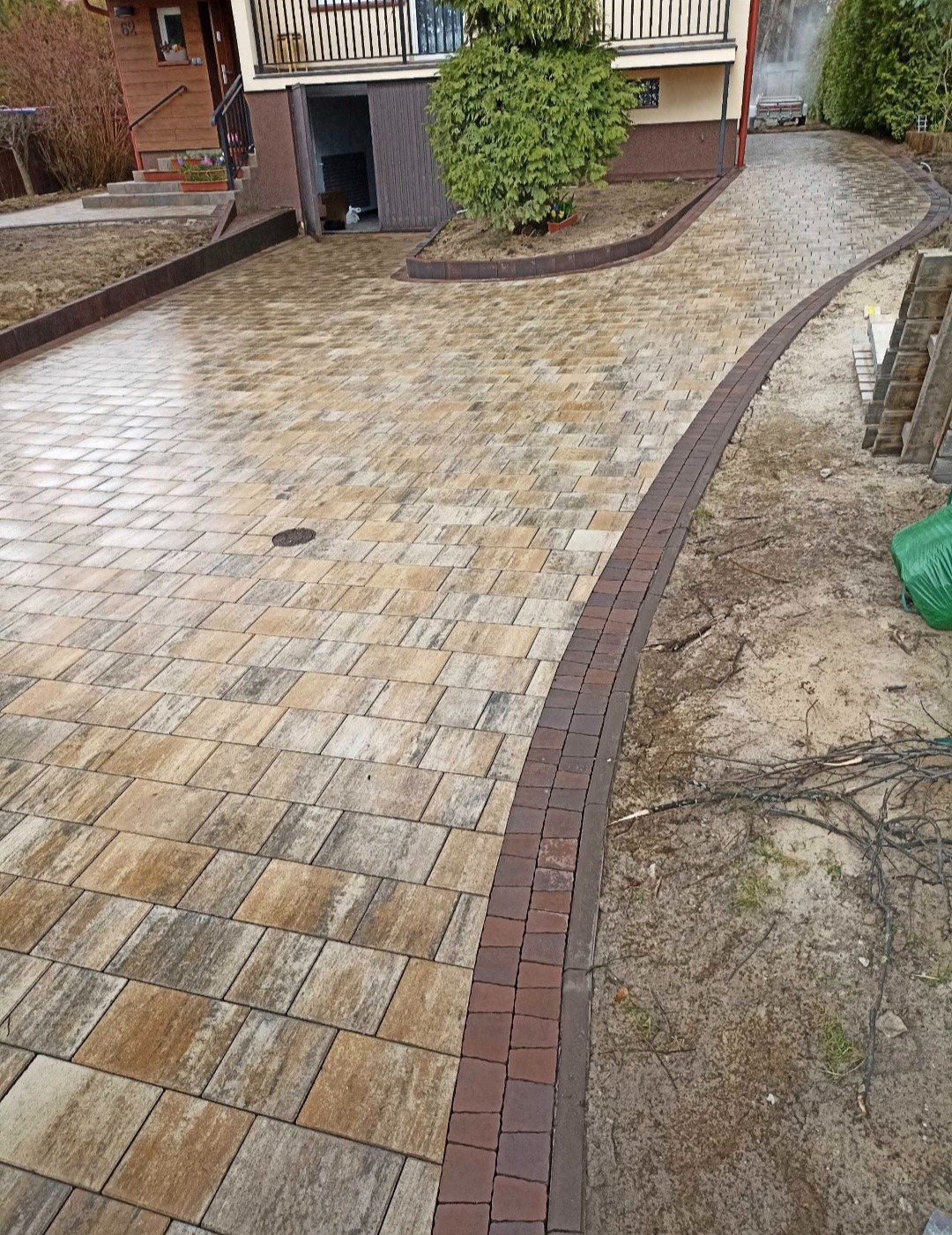 Ułożona, wilgotna nawierzchnia z kostki brukowej w odcieniach beżu i brązu, obrzeże z ciemniejszej kostki, widok na dom z zielonym krzewem i fragmentem ogrodu.