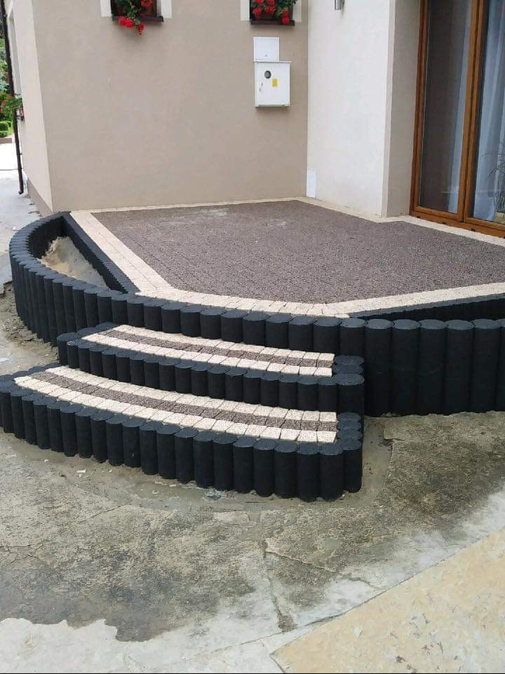 Ułożona nawierzchnia z kostki brukowej imitującej drewniane bale, tworząca schody i taras przed domem z kwiatami w oknach.