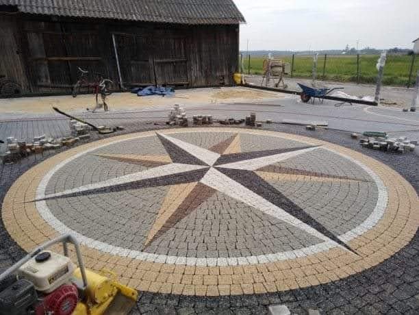 Okrąg z kostki brukowej ułożony w kształt kompasu z gwiazdą w centrum, otoczony obwódką z żółtej kostki, w tle budynek gospodarczy i narzędzia budowlane.