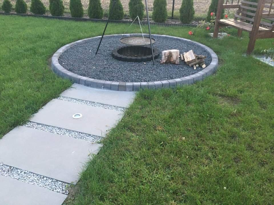 Ogród z okrągłym paleniskiem otoczonym kostką, ścieżka z płyt z wbudowanym oświetleniem i dekoracyjnym żwirem, w tle tuje i drewniana ławka.
