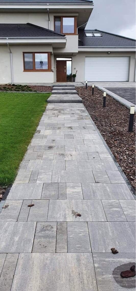 Ułożona ścieżka z szarej kostki brukowej prowadząca do domu z jasną elewacją, widoczne schody z kostki, trawnik i rabata z kory.