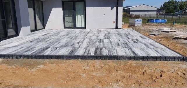 Ułożona szaro-biała kostka brukowa przed nowoczesnym domem z czarnymi ramami okiennymi, obrzeża z ciemnej kostki, teren wokół w trakcie przygotowania.