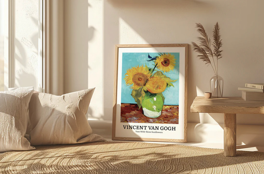 Wydruk oprawionego plakatu z reprodukcją obrazu Vincenta van Gogha 'Wazon z trzema słonecznikami' ustawionego na podłodze obok beżowych poduszek i stolika z dekoracjami w jasnym wnętrzu.