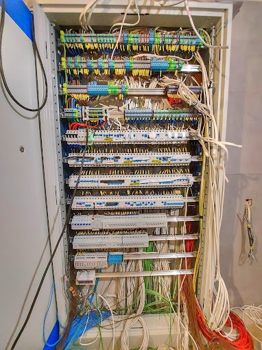 Otwarta szafa elektryczna z widocznymi bezpiecznikami, przewodami w różnych kolorach i złączkami, częściowo zamontowana, z widocznymi przewodami zwisającymi na zewnątrz.