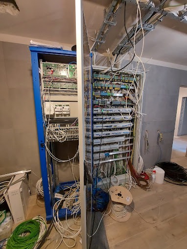 Rozdzielnica elektryczna w trakcie montażu, widoczne liczne kable, przewody w różnych kolorach, skrzynka elektryczna w kolorze niebieskim, wnętrze w stanie surowym.