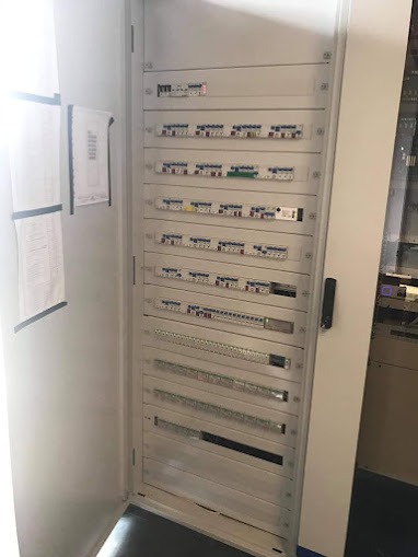 Otwarta szafa elektryczna z wieloma bezpiecznikami i listwami zaciskowymi, widoczne dokumenty przywieszone po lewej stronie.