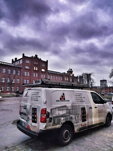 Biały samochód dostawczy firmy Urban Technology System z drabiną na dachu, zaparkowany przed ceglanym budynkiem na tle pochmurnego nieba. Na karoserii widoczne grafiki przedstawiające systemy...