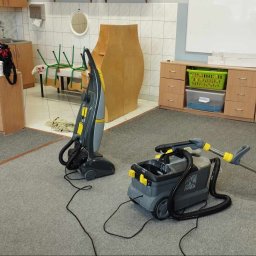 SzuruSzuru.pl - Dwa profesjonalne odkurzacze piorące na szarym dywanie w pomieszczeniu z meblami biurowymi i białymi ścianami. Jeden odkurzacz pionowy, drugi z pojemnikiem na wodę i wężem. Widoczny fragment...