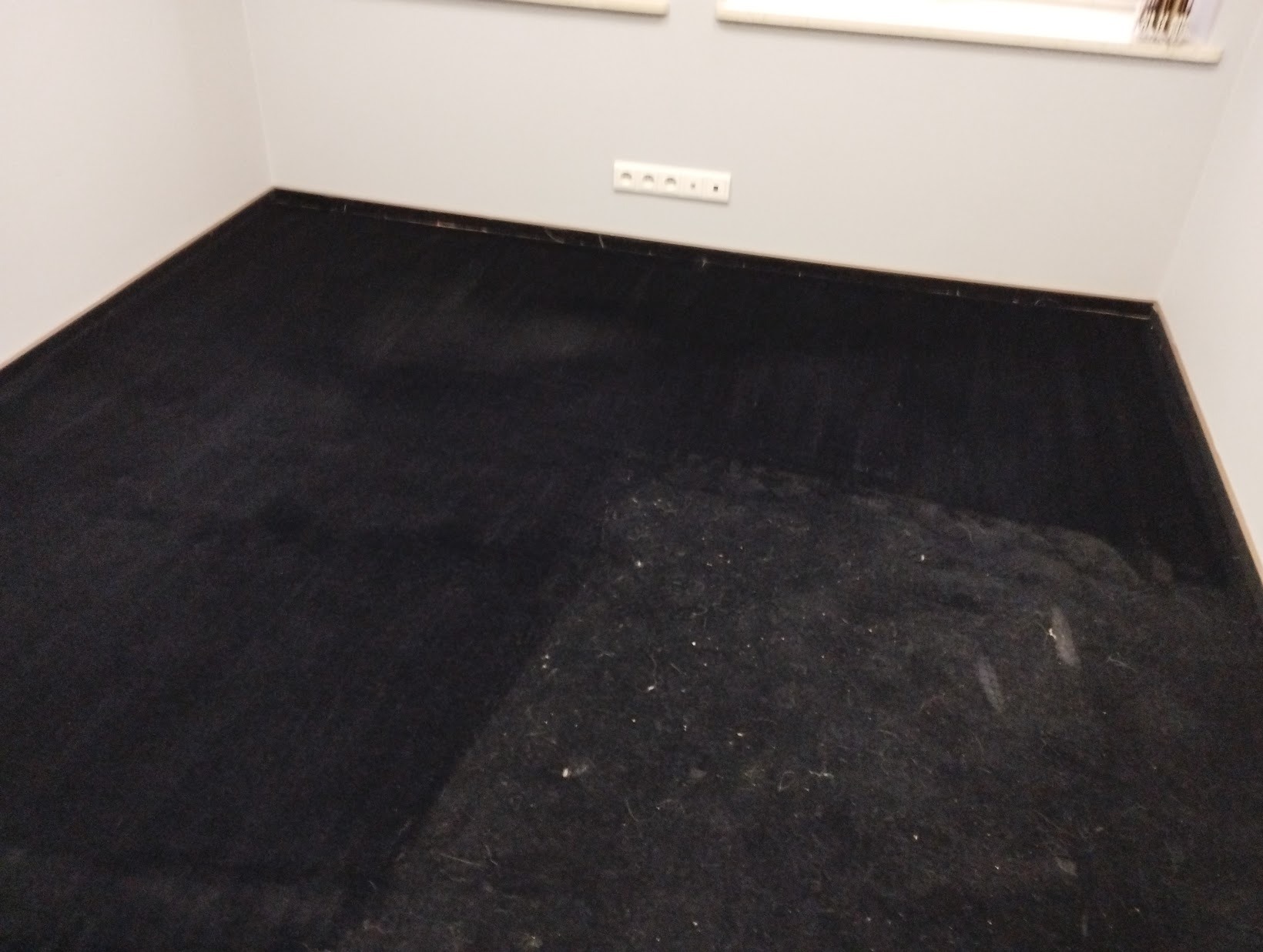 Ciemny dywan po czyszczeniu, widoczne jaśniejsze plamy i drobne zanieczyszczenia, ściana z gniazdkiem elektrycznym w tle.