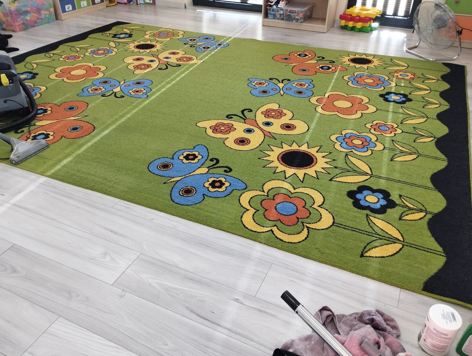 Czyszczenie zielonego dywanu dziecięcego z motywem kwiatów i motyli za pomocą odkurzacza piorącego; widoczna końcówka odkurzacza i fragment urządzenia, jasne panele podłogowe, zabawki w tle.