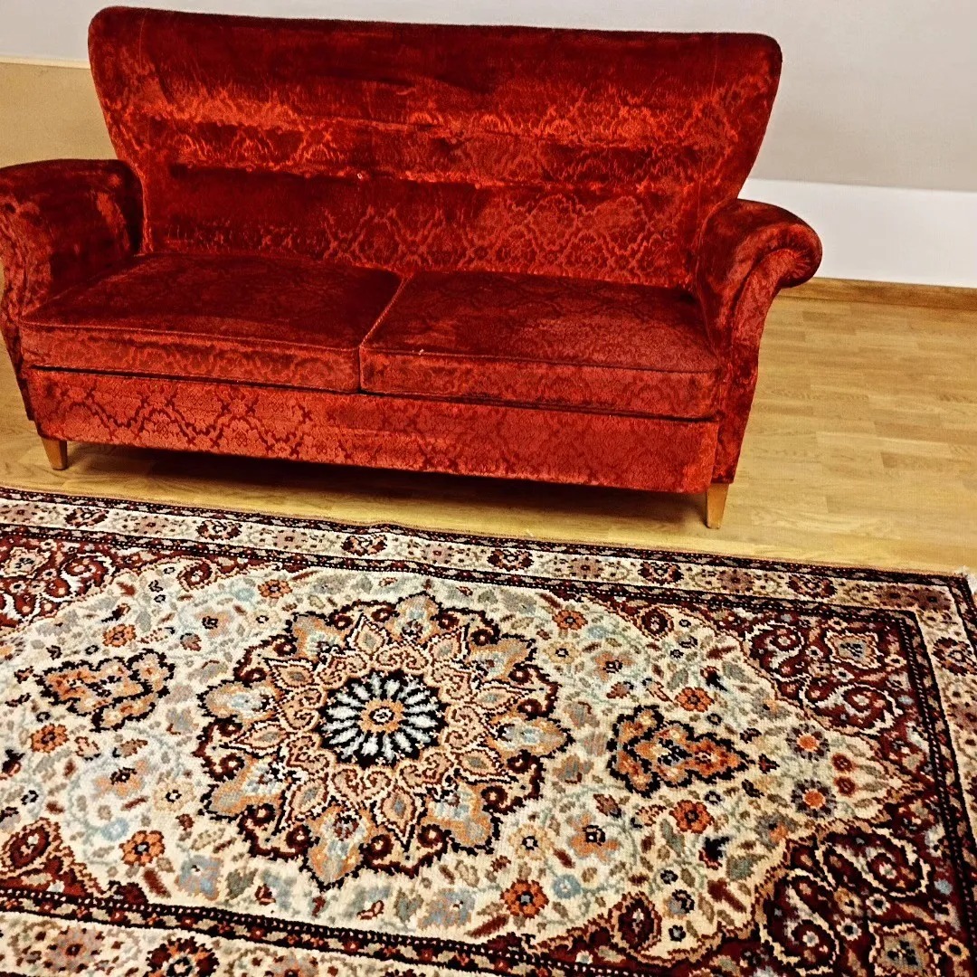 Czerwona, welurowa sofa z wysokim oparciem stoi na drewnianej podłodze obok wzorzystego dywanu orientalnego.