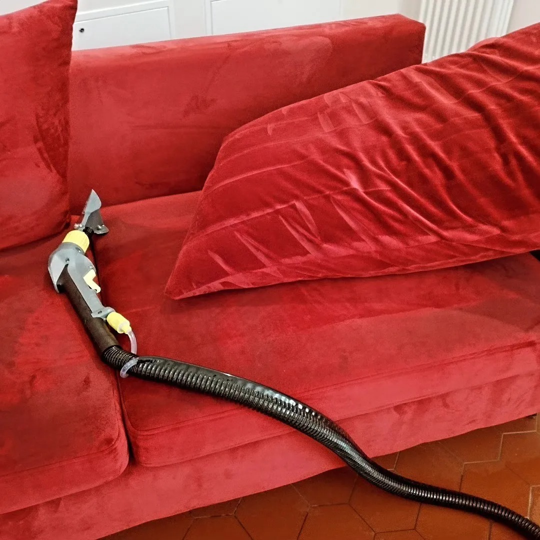 Czerwona welurowa kanapa podczas czyszczenia parowego, widoczny fragment czyszczonej powierzchni i dysza urządzenia z czarnym wężem.