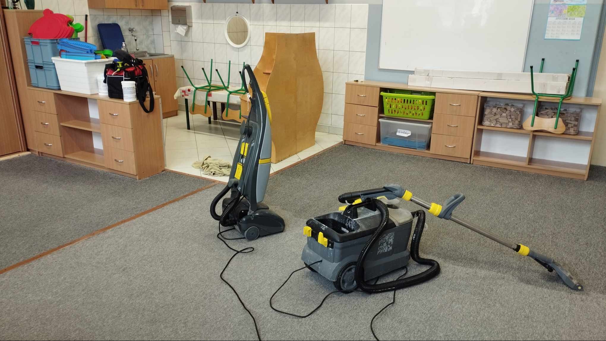 Dwa profesjonalne odkurzacze piorące na szarym dywanie w pomieszczeniu z meblami biurowymi i białymi ścianami. Jeden odkurzacz pionowy, drugi z pojemnikiem na wodę i wężem. Widoczny fragment...