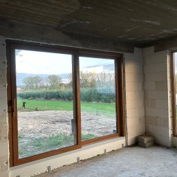 TOMEX OKNA SPÓŁKA Z OGRANICZONĄ ODPOWIEDZIALNOŚCIĄ - Montaż brązowych drzwi przesuwnych w surowym stanie, widok na zewnątrz z osobą na trawniku i budynkiem w tle.