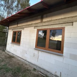 TOMEX OKNA SPÓŁKA Z OGRANICZONĄ ODPOWIEDZIALNOŚCIĄ - Nowo zamontowane okna z brązowymi ramami w surowym budynku z białych bloczków, widoczne ślady zaprawy i niezakończone prace budowlane.