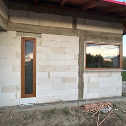 TOMEX OKNA SPÓŁKA Z OGRANICZONĄ ODPOWIEDZIALNOŚCIĄ - Surowy stan budynku z zamontowanymi oknami z brązowymi ramami, uszczelnionymi pianką montażową, widoczne bloczki betonowe i drewniane elementy szalunku.