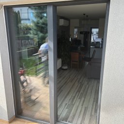 TOMEX OKNA SPÓŁKA Z OGRANICZONĄ ODPOWIEDZIALNOŚCIĄ - Nowoczesne, szare drzwi przesuwne z widokiem na wnętrze domu z drewnianą podłogą i kuchnią w tle oraz na zewnątrz z widocznym ogrodem i narzędziami malarskimi.