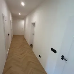 Korytarz z jasną podłogą ułożoną we wzór jodełki, białymi ścianami i białymi drzwiami z czarnymi klamkami; widoczne czarne kontakty na ścianie.