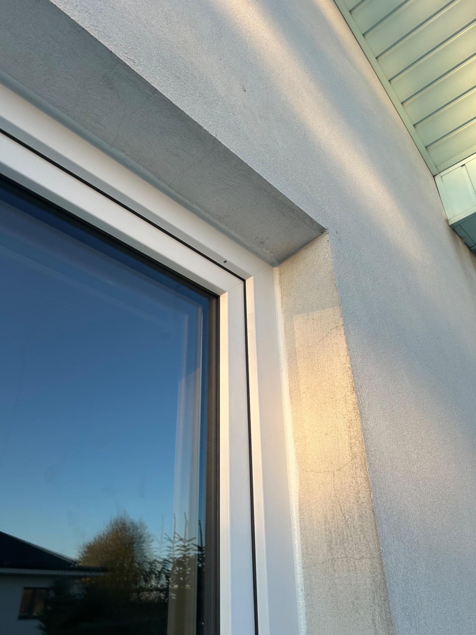 Fragment białej ramy okiennej z widocznym odbiciem nieba i drzew na tle szarego tynku elewacyjnego, oświetlonego popołudniowym słońcem.