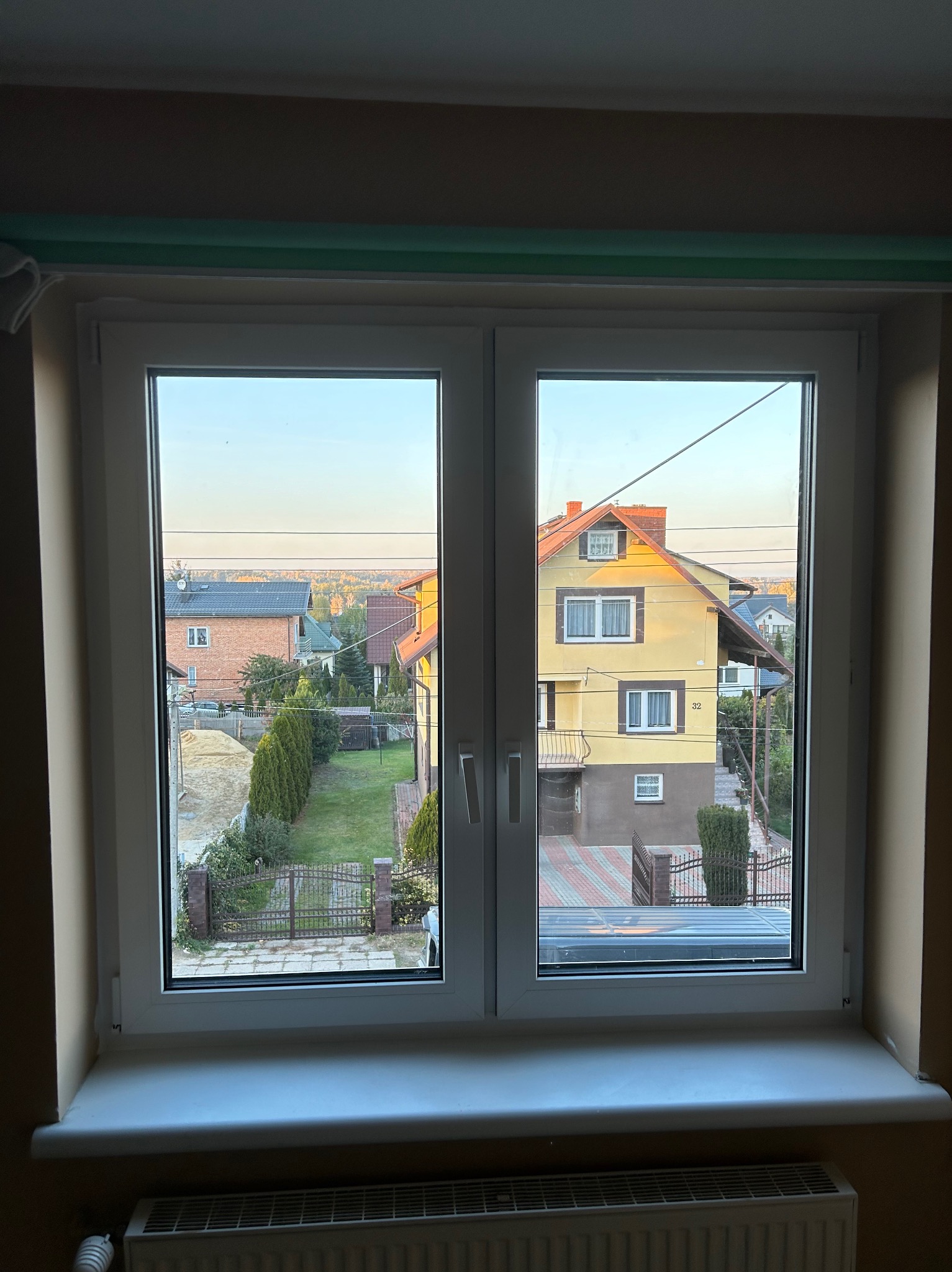 Dwuskrzydłowe białe okno z widokiem na podwórko z domami w oddali, wczesny poranek.