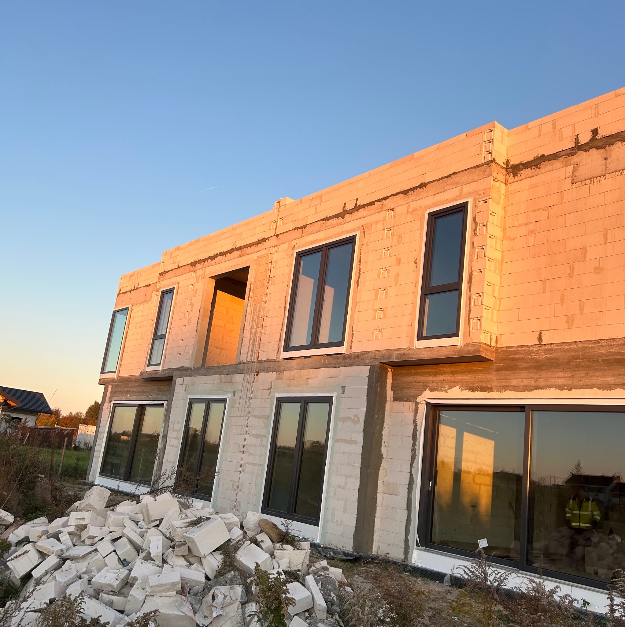 Budynek w trakcie budowy z zamontowanymi oknami z czarnymi ramami, widoczny stos białych bloczków budowlanych, ciepłe światło zachodzącego słońca.