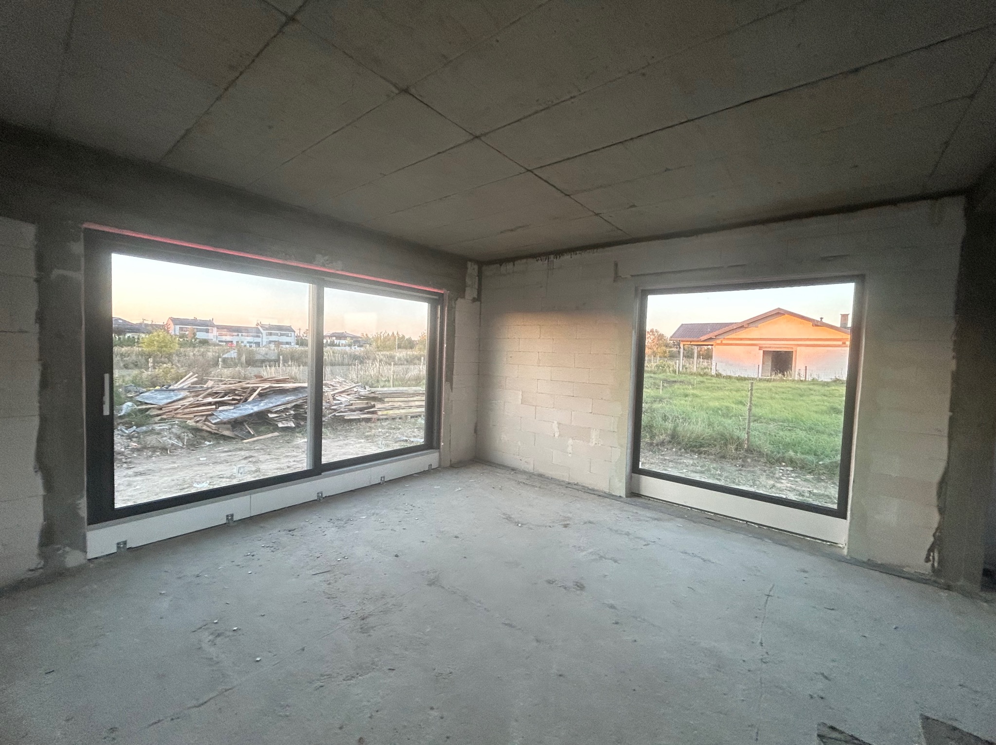 Surowe wnętrze w trakcie budowy z zamontowanymi dużymi oknami z czarnymi ramami, widok na zewnątrz na plac budowy i zachodzące słońce.