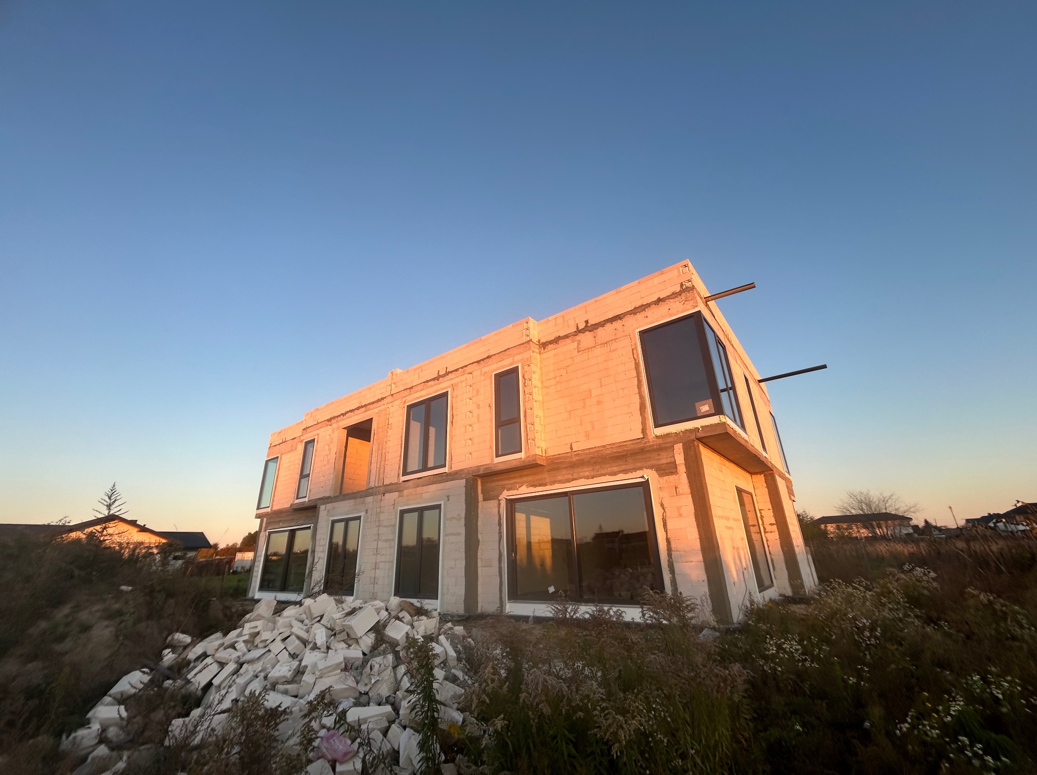 Surowy stan dwukondygnacyjnego domu z pustaków z zamontowanymi oknami, widoczne pozostałości materiałów budowlanych i zachodzące słońce.