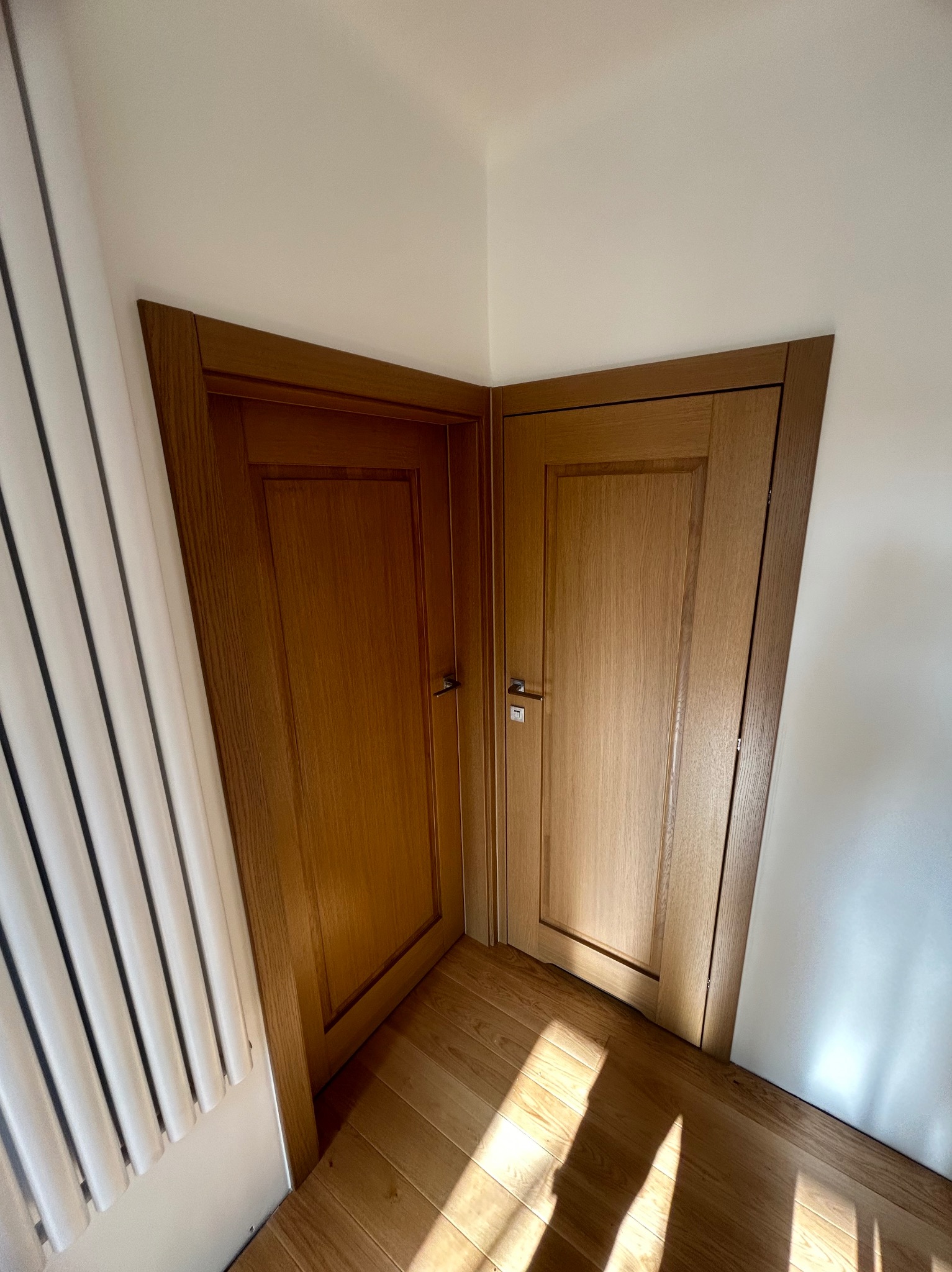 Dwa drewniane drzwi wewnętrzne, ustawione pod kątem w rogu pomieszczenia, z widocznym fragmentem grzejnika i parkietem w ciepłym odcieniu, oświetlone naturalnym światłem.