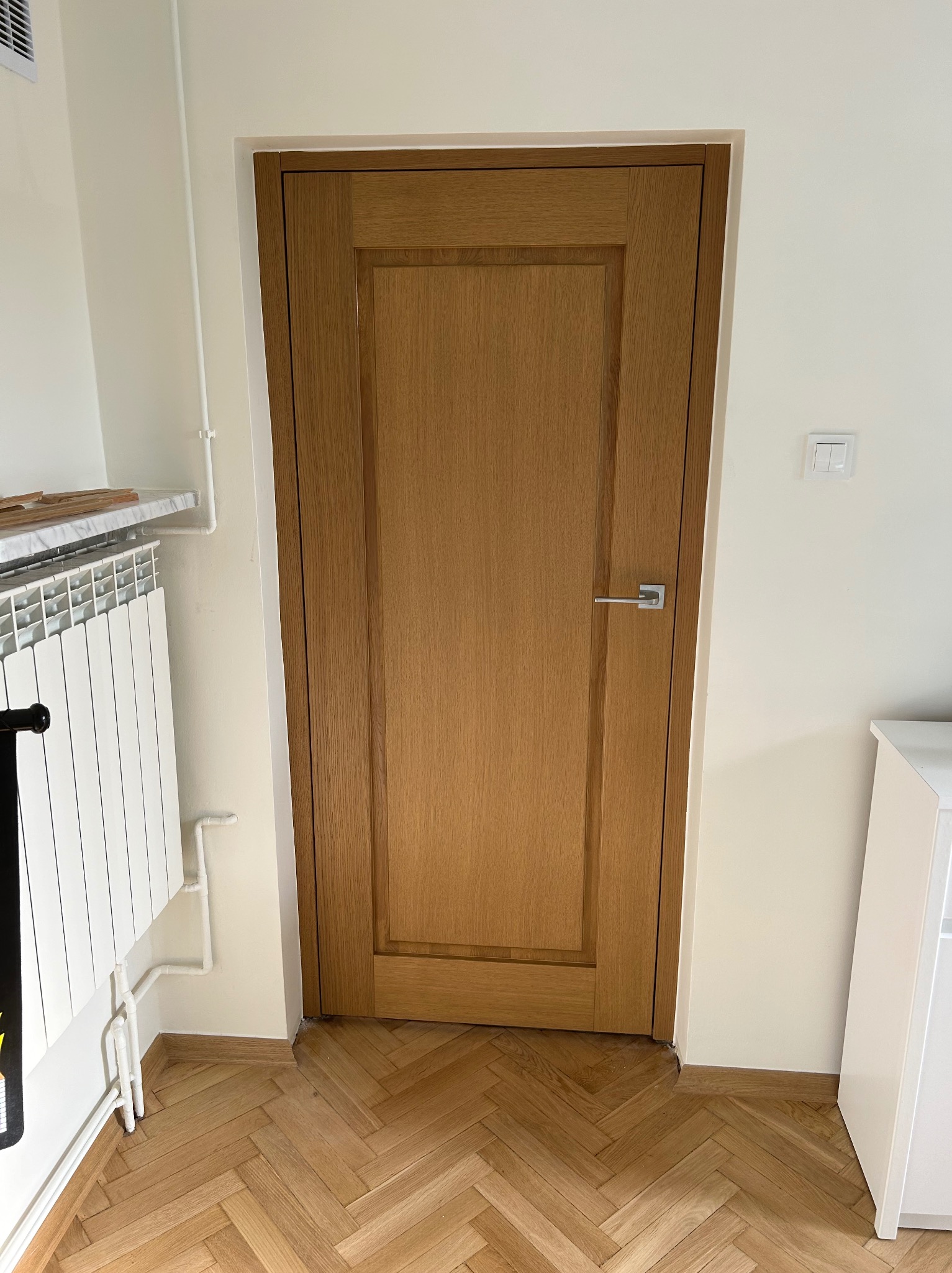 Dębowa, ramowa konstrukcja drzwi wewnętrznych z klamką, widoczna na tle parkietu ułożonego w jodełkę i białych ścian.