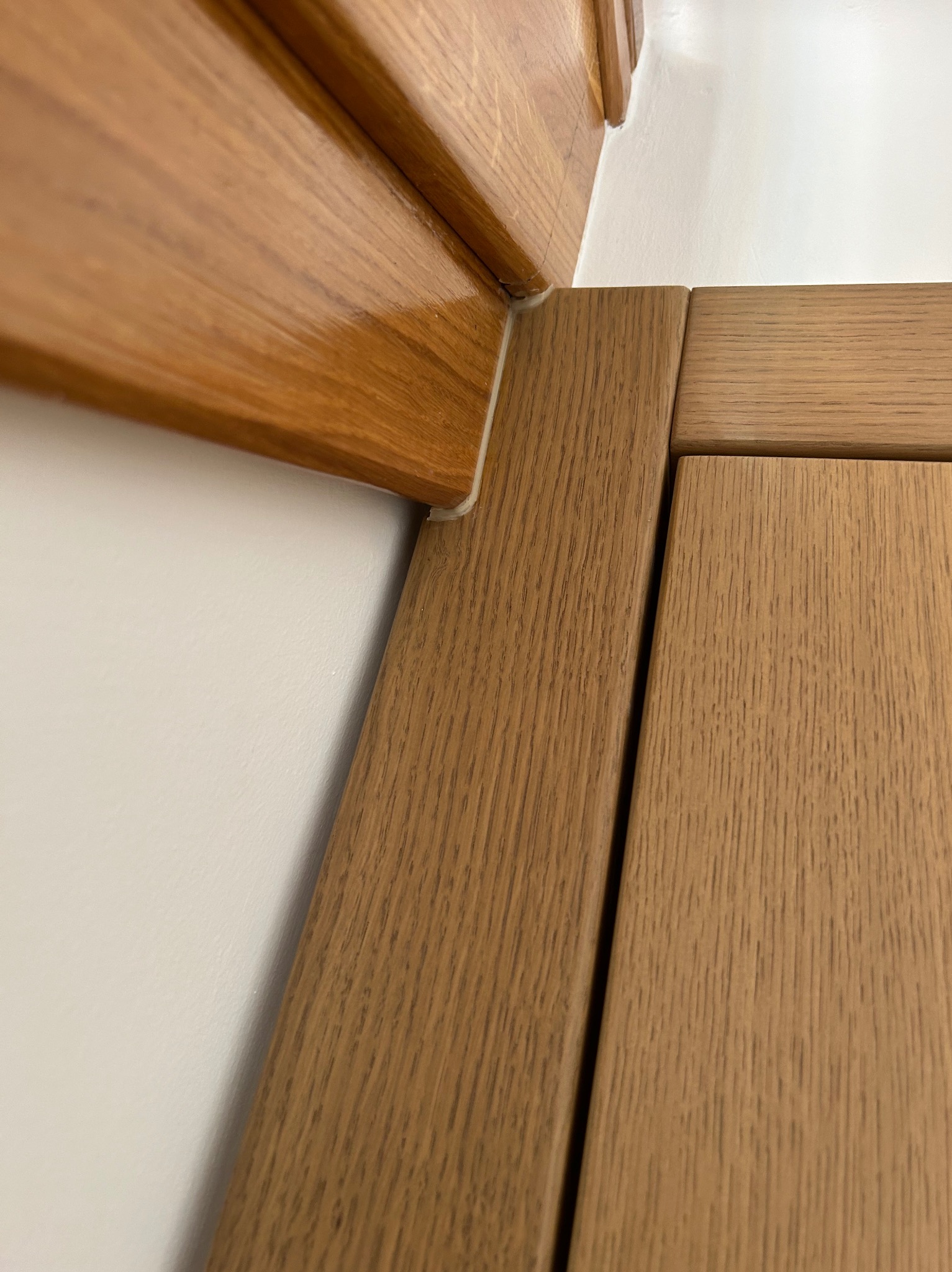 Detal drewnianej ościeżnicy i fragmentu skrzydła drzwiowego w odcieniach brązu, widoczne wypełnienie silikonowe w narożniku, na tle białej ściany.