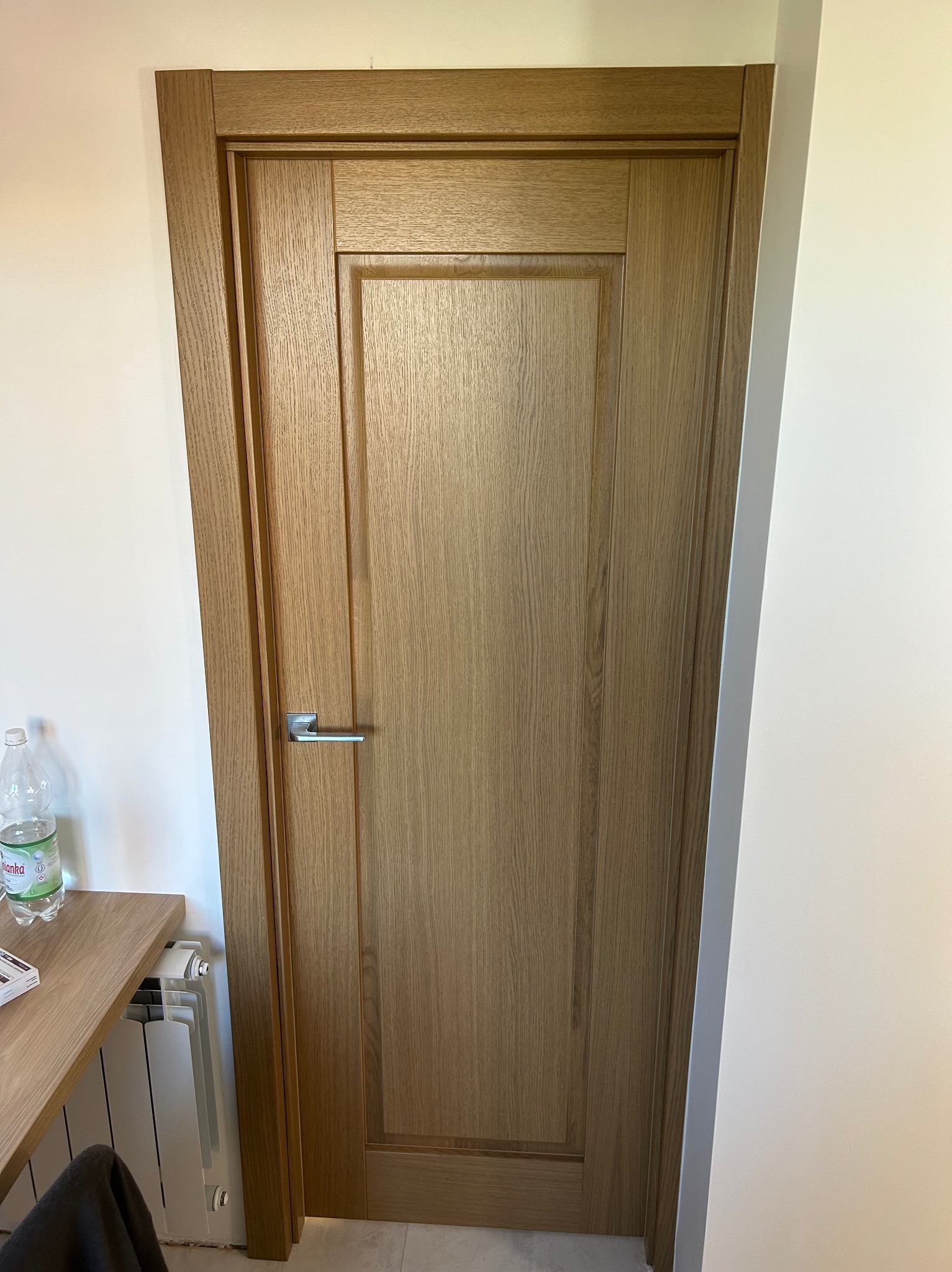 Dębowe drzwi wewnętrzne z prostokątnym panelem i srebrną klamką, widoczne w jasnym wnętrzu obok białego grzejnika i fragmentu drewnianego blatu.