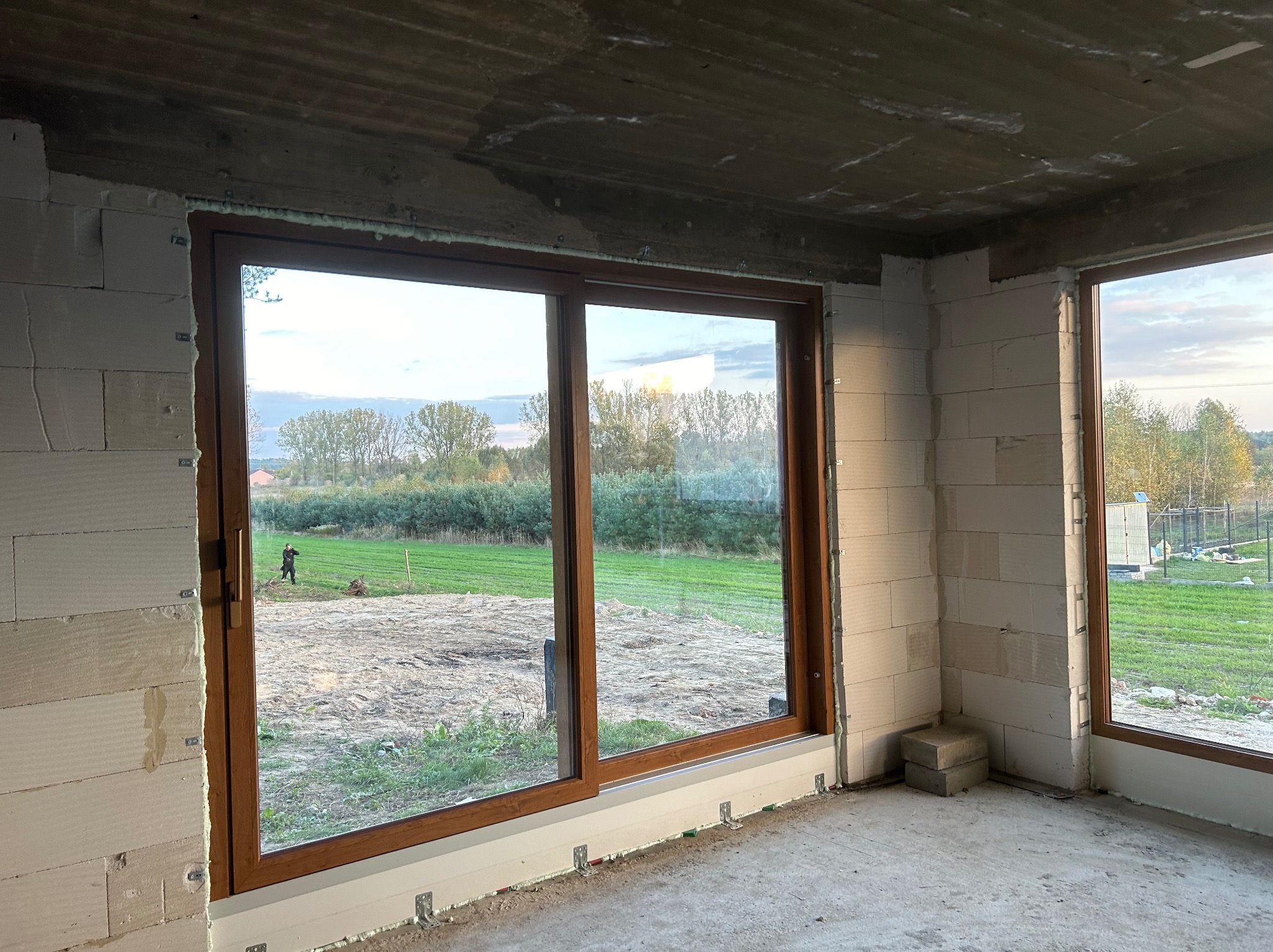 Montaż brązowych drzwi przesuwnych w surowym stanie, widok na zewnątrz z osobą na trawniku i budynkiem w tle.