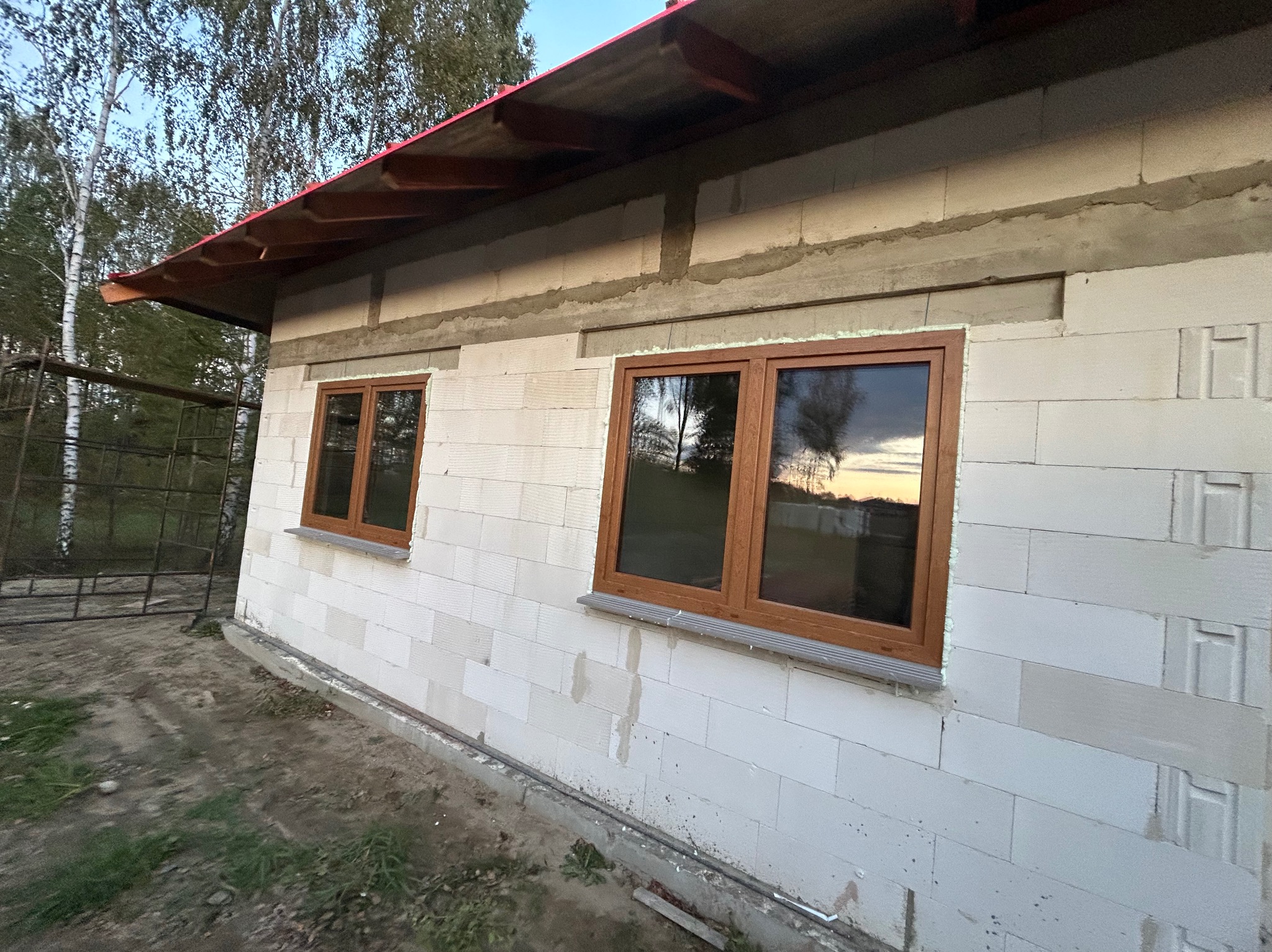 Nowo zamontowane okna z brązowymi ramami w surowym budynku z białych bloczków, widoczne ślady zaprawy i niezakończone prace budowlane.