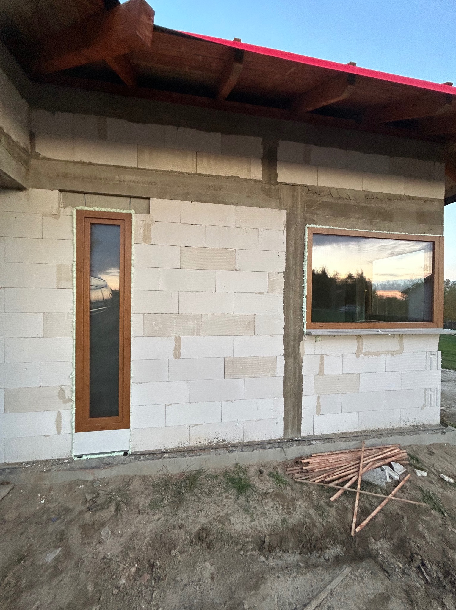 Surowy stan budynku z zamontowanymi oknami z brązowymi ramami, uszczelnionymi pianką montażową, widoczne bloczki betonowe i drewniane elementy szalunku.