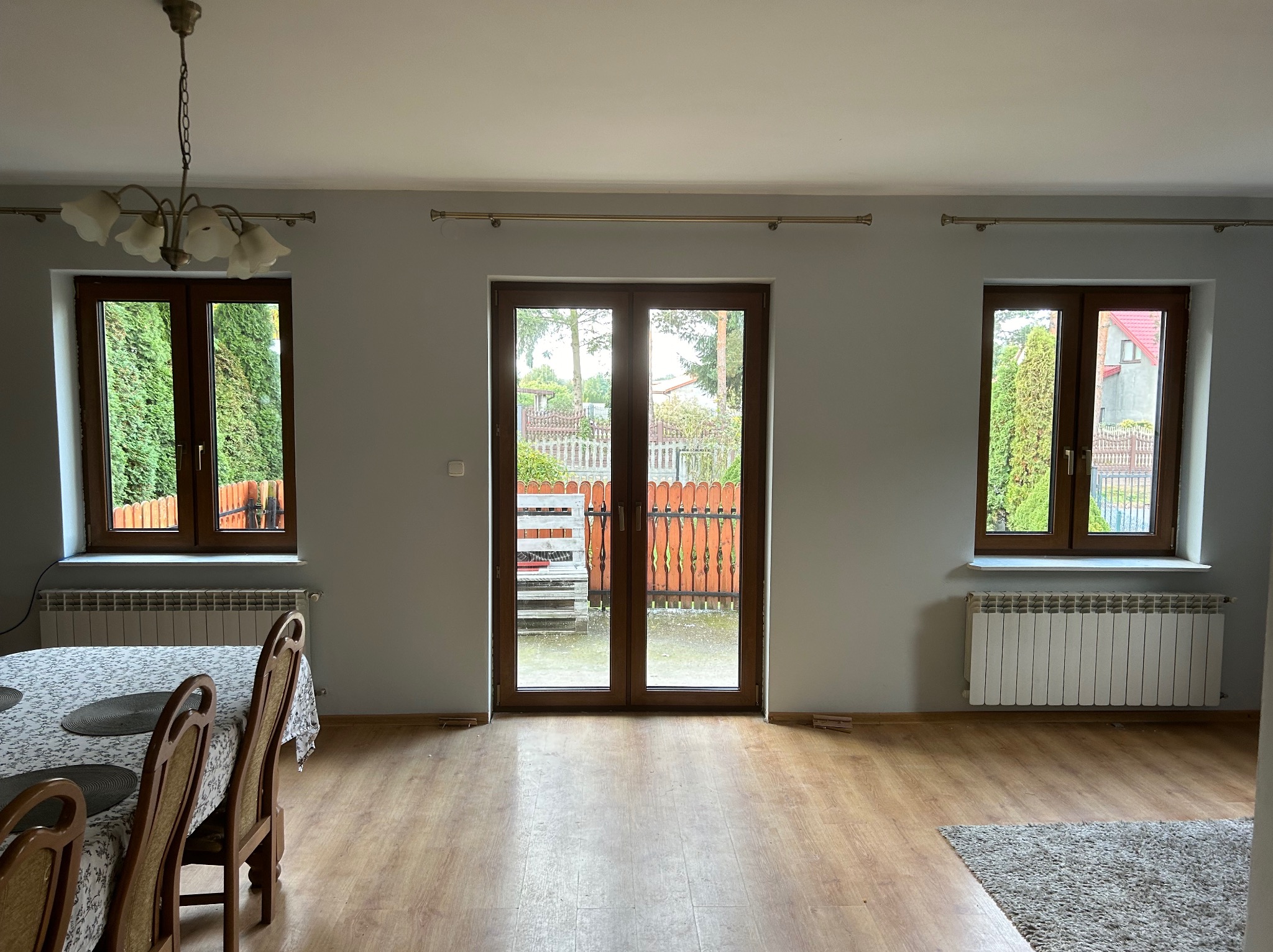 Wnętrze pokoju z drewnianymi oknami PCV i drzwiami balkonowymi, widok na ogród przez okna, grzejnik pod oknem, stół z krzesłami w rogu pokoju.